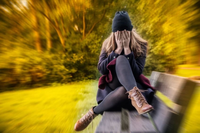 woman sitting on bench outside covering face with hands, surrounded by trees filled with anxiety